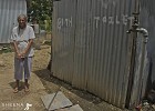 Louise Yearwood in her back yard .jpg - Louise in back yard. The shower and toilet are enclosed by a corrugated iron structure. The outside tap is also used for washing.